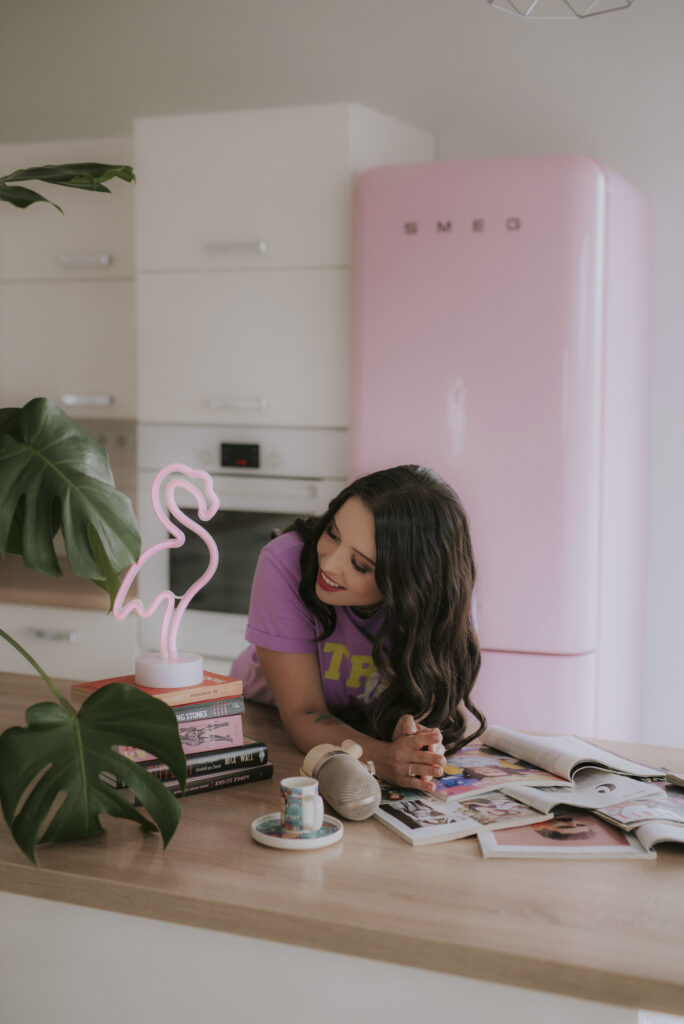 Autorka Astrokultury Agata pozująca na tle różowej lodówki Smeg, oparta o blat, z książkami i prasą w tle